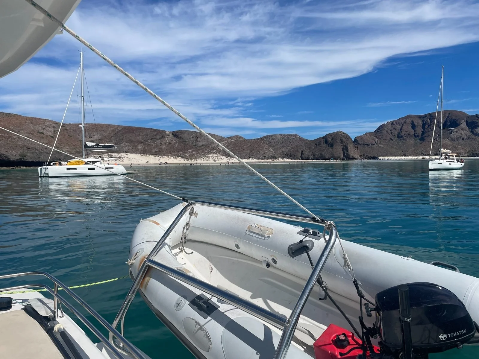 View from a boat of a calm bay with two sailboats anchored, surrounded by brown rocky hills, and partly cloudy blue sky.