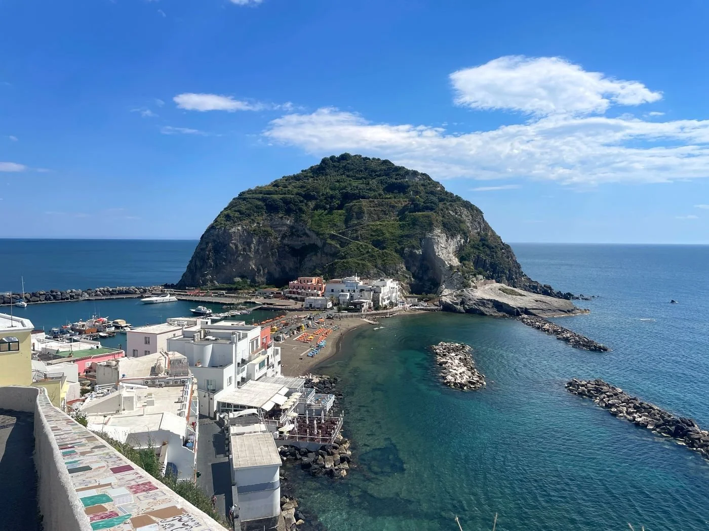 A scenic coastal view featuring a small harbor with boats, white buildings along the shore, and a forested mountain in the background under a clear blue sky with scattered clouds.