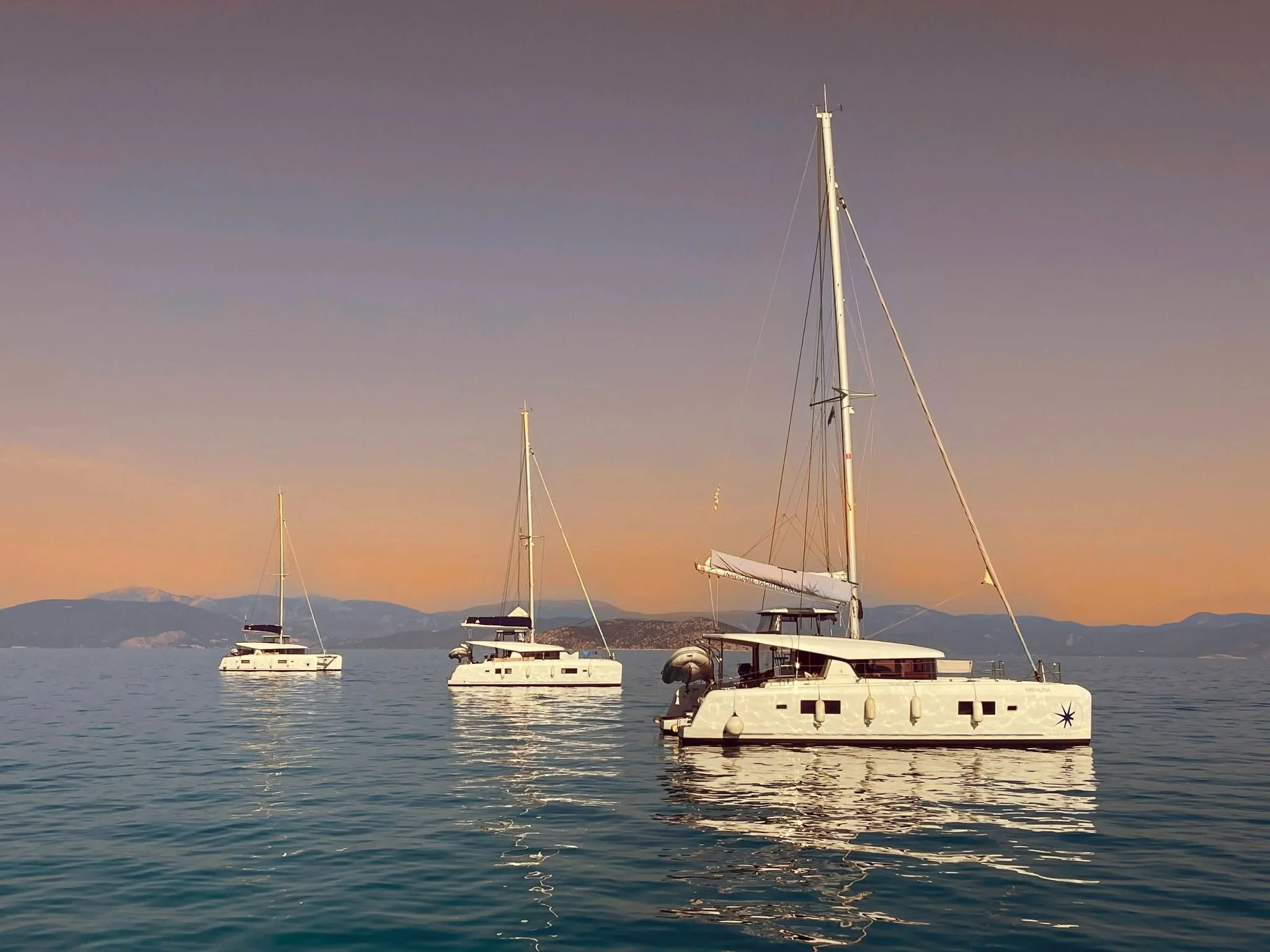 Several yachts docked on a calm body of water during sunset, with mountain ranges in the background and a clear sky with pastel colors.