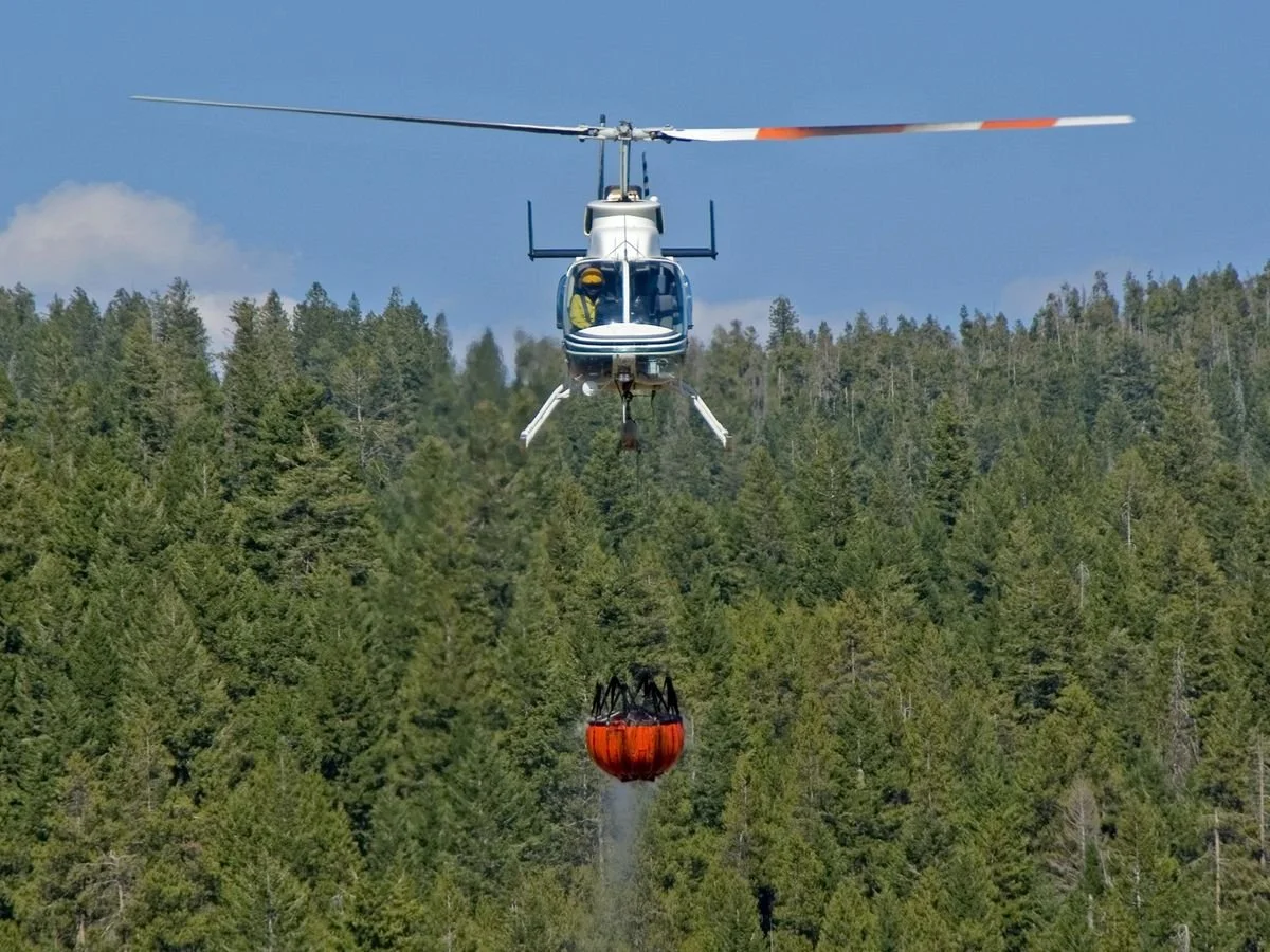 A helicopter hovering over a forested area, releasing a bucket of water for firefighting.