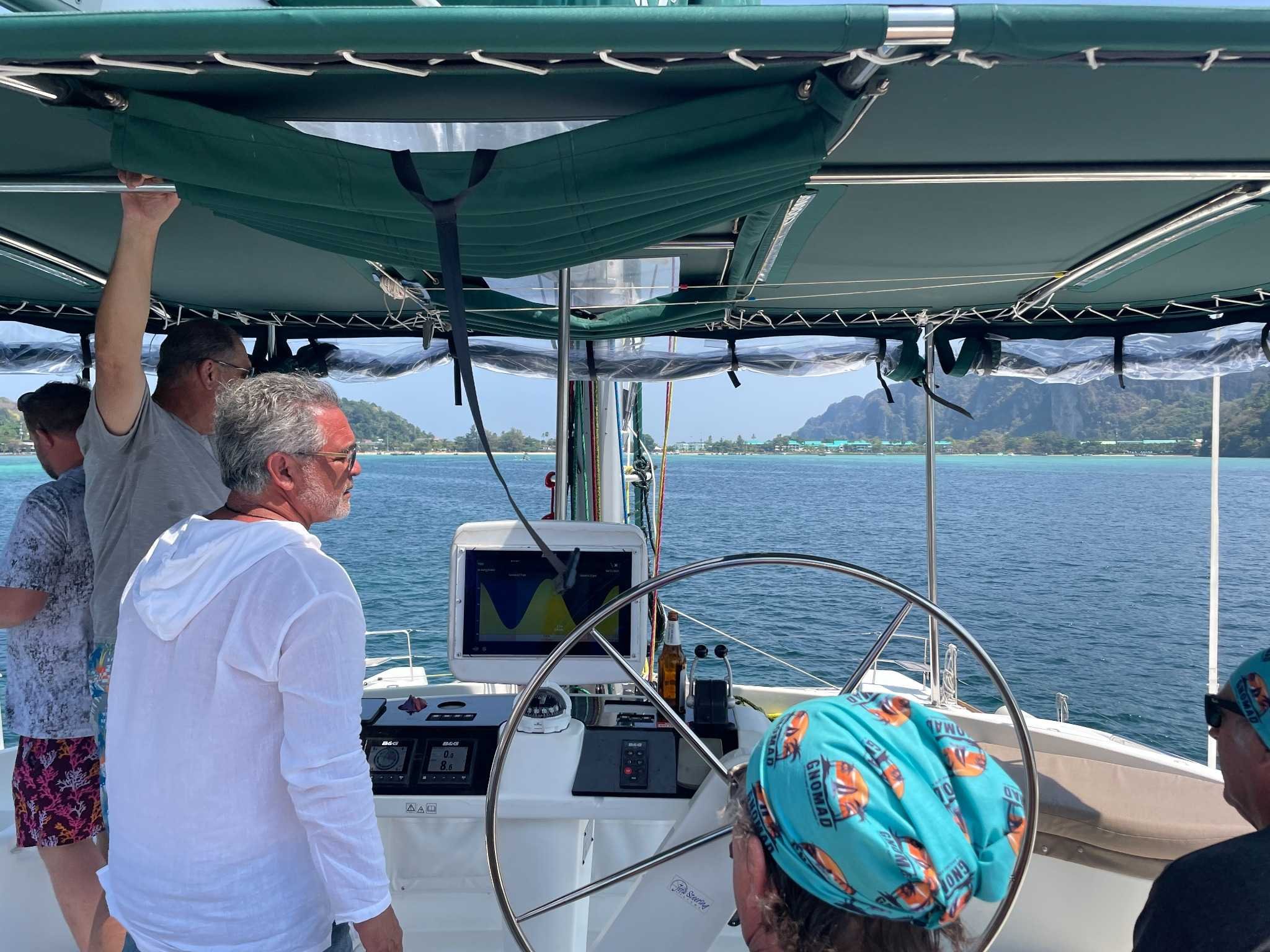 People on a boat, with a mountainous coastline and blue water in the background, and a radar or navigation display in the center of the boat.