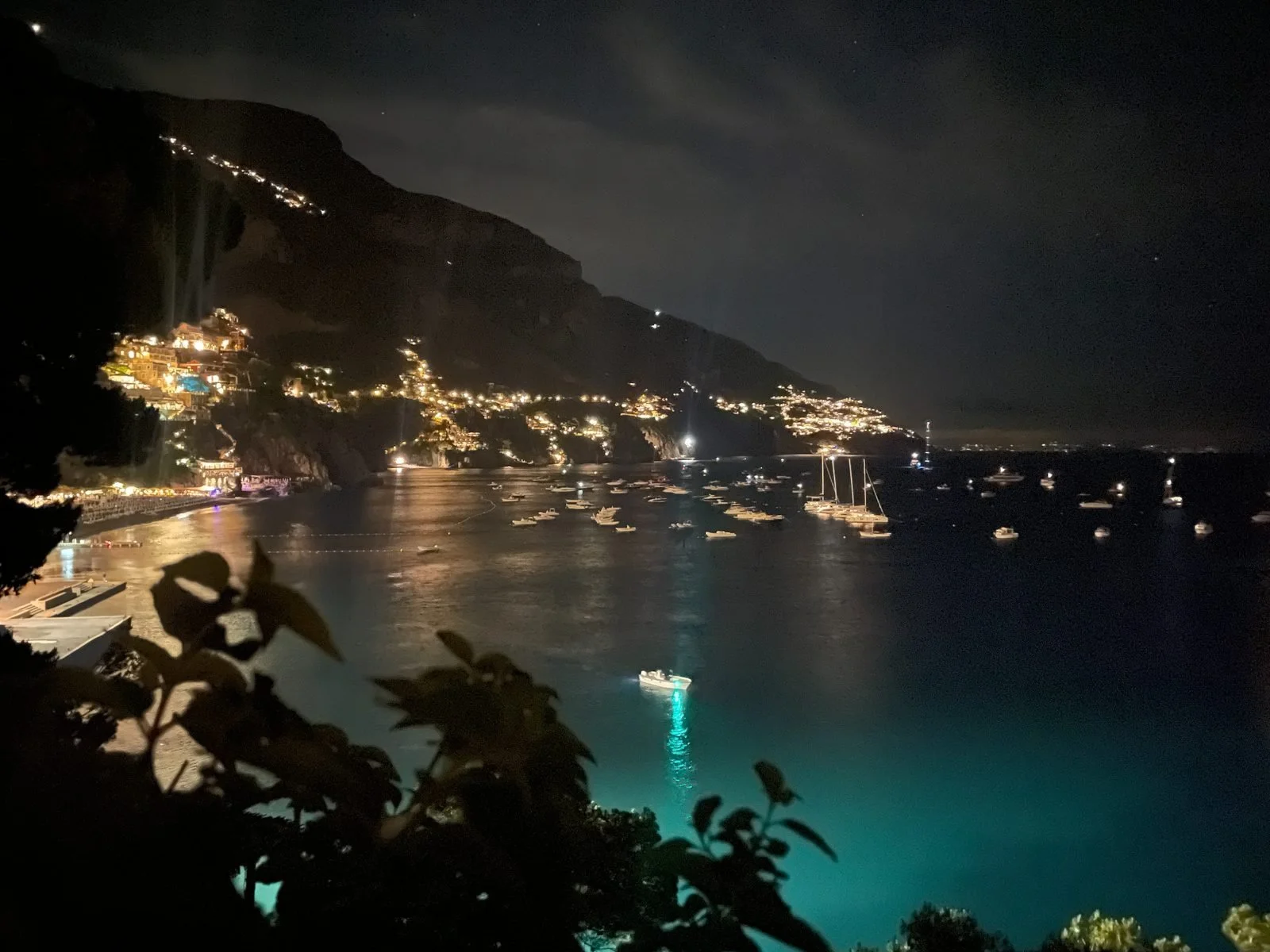 Nighttime view of a coastal town with lights along the hillside, boats in the water, and reflections on the ocean.