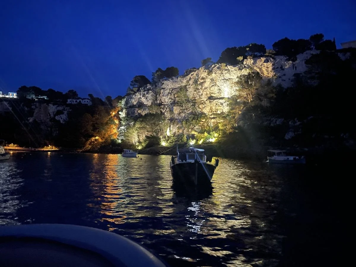 Nighttime view of a boat on calm water with illuminated rocky shoreline and trees in the background under a dark blue sky.