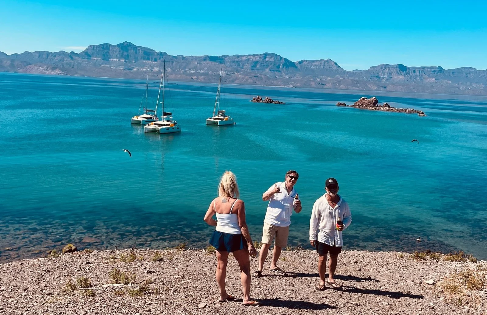 Three people standing on a rocky shore by a bright blue lake with sailboats in the water and mountains in the background, enjoying sunny weather.