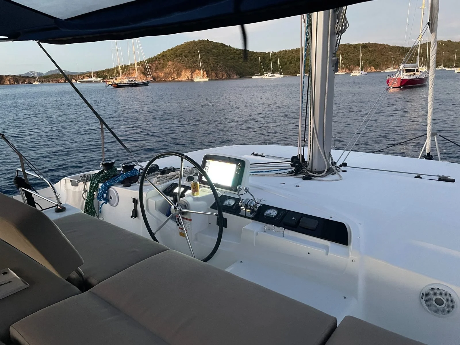 View from inside a sailing boat showing the steering wheel, navigation equipment, and the deck with surrounding water and other boats anchored nearby.