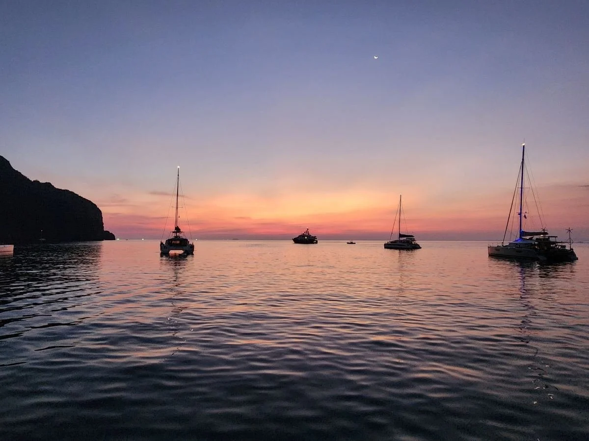 Boats floating on calm water during sunset, with a colorful sky and crescent moon overhead.