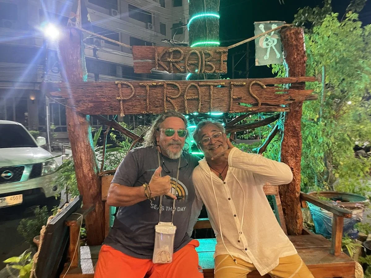 Two men posing in front of a wooden sign that says 'KROB Pirates' at night, with one giving a thumbs-up and the other making a gesture near his face, in front of a pirate-themed structure with green lights.