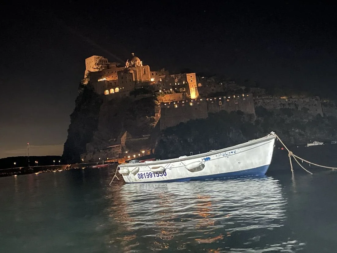 A boat floating in calm water at night with a lit castle on a cliff in the background.