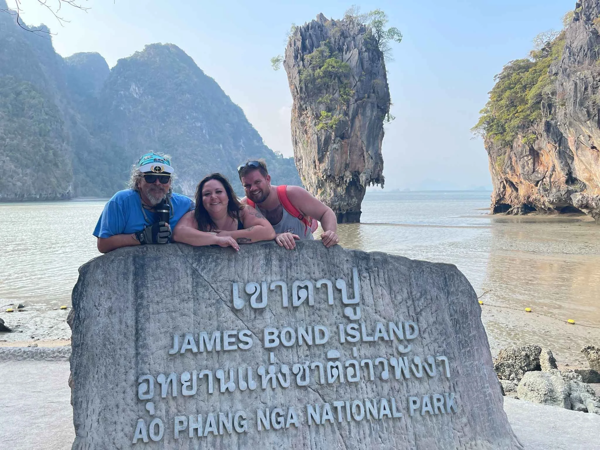 Three people posing behind a large rock with a sign reading 'James Bond Island' in a body of water surrounded by tall limestone cliffs and a clear sky.