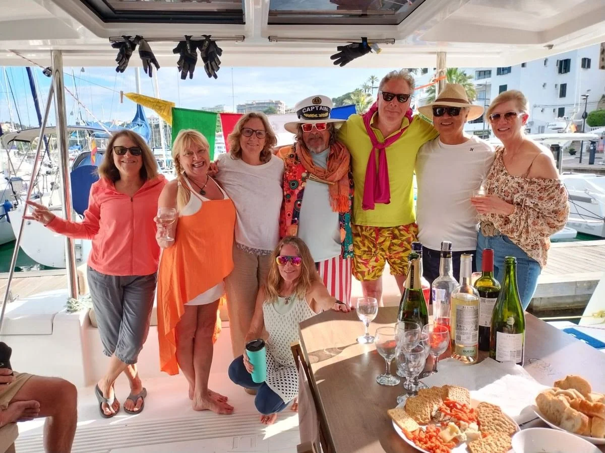 A group of nine people on a boat docked at a marina, celebrating with food and drinks, dressed in colorful casual summer clothing and accessories, with a table of snacks and beverages in front.
