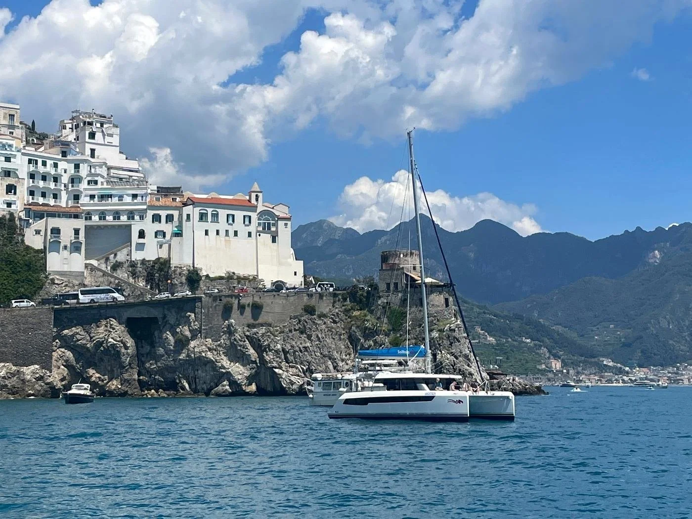 A scenic view of a coast with white buildings on a hillside, a sailboat in the water, and mountains in the background under a blue sky with clouds.