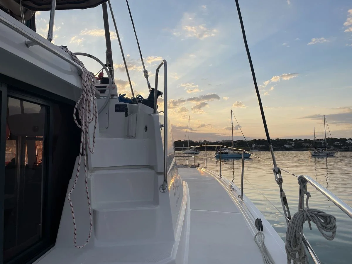 View from a boat's deck at sunset, showing calm water, several anchored sailboats, and a distant shoreline with houses.