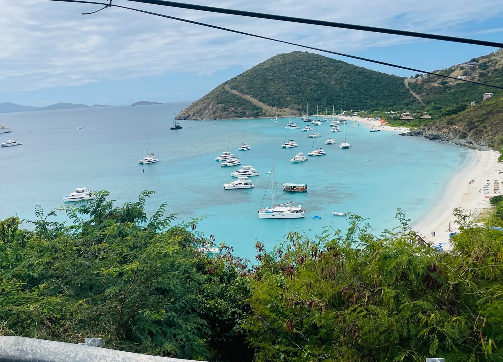 A scenic view of a bay with numerous sailboats anchored in clear blue water, a sandy beach on the right, a hillside covered with green trees, and cloudy sky.