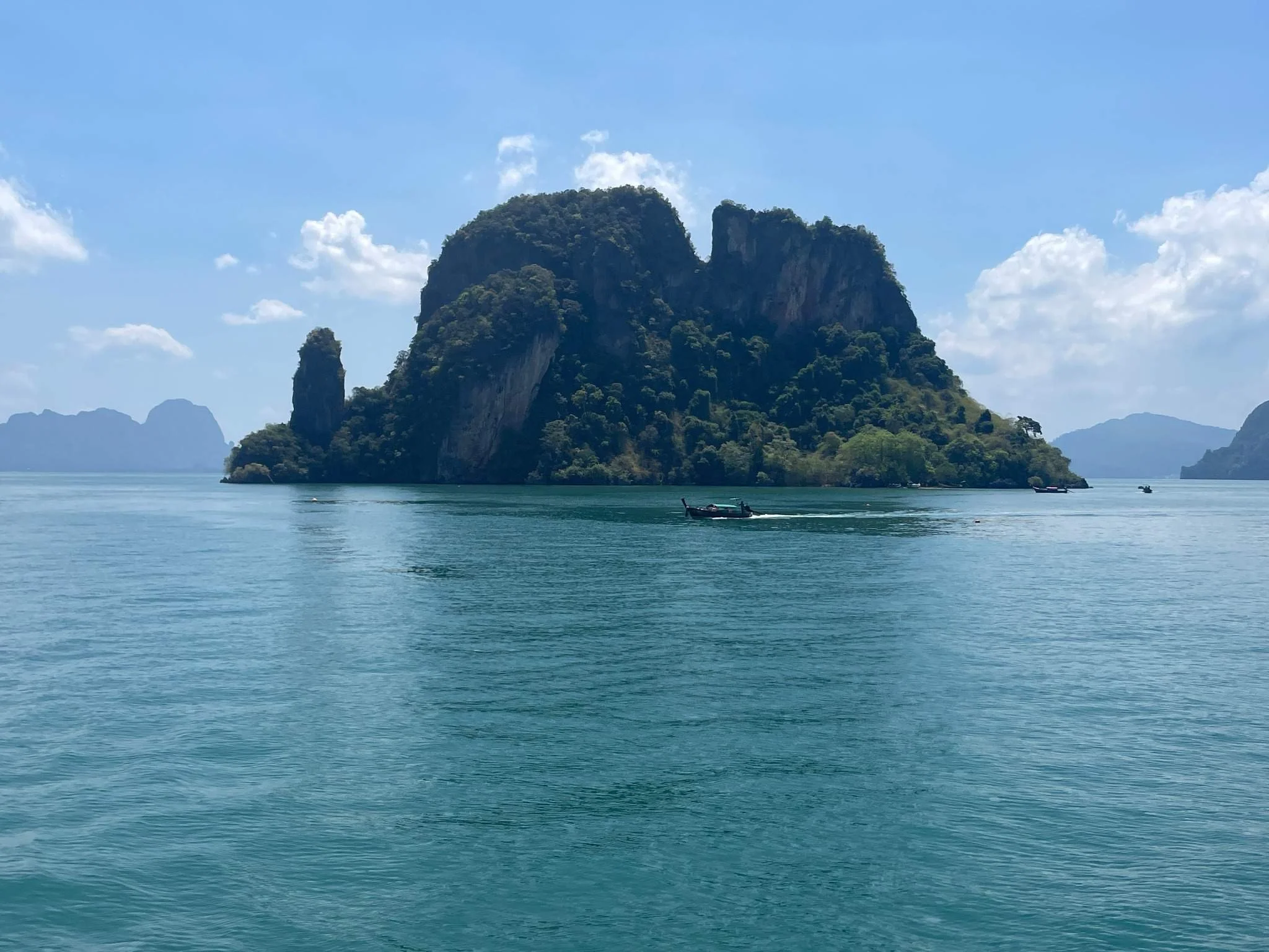 A large green island with steep cliffs rising from turquoise water, with a few boats floating nearby, under a partly cloudy sky.