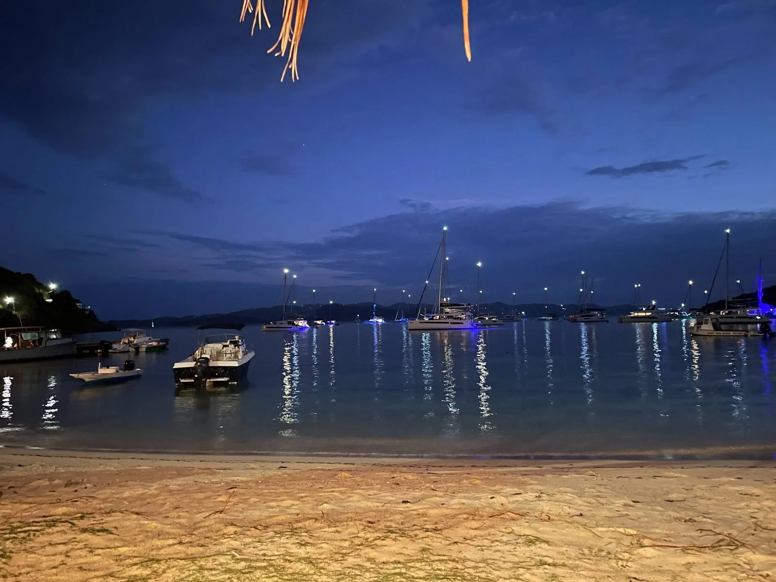 Boats docked at a marina during nighttime, with illuminated lights reflecting on calm water, under a dark blue sky.