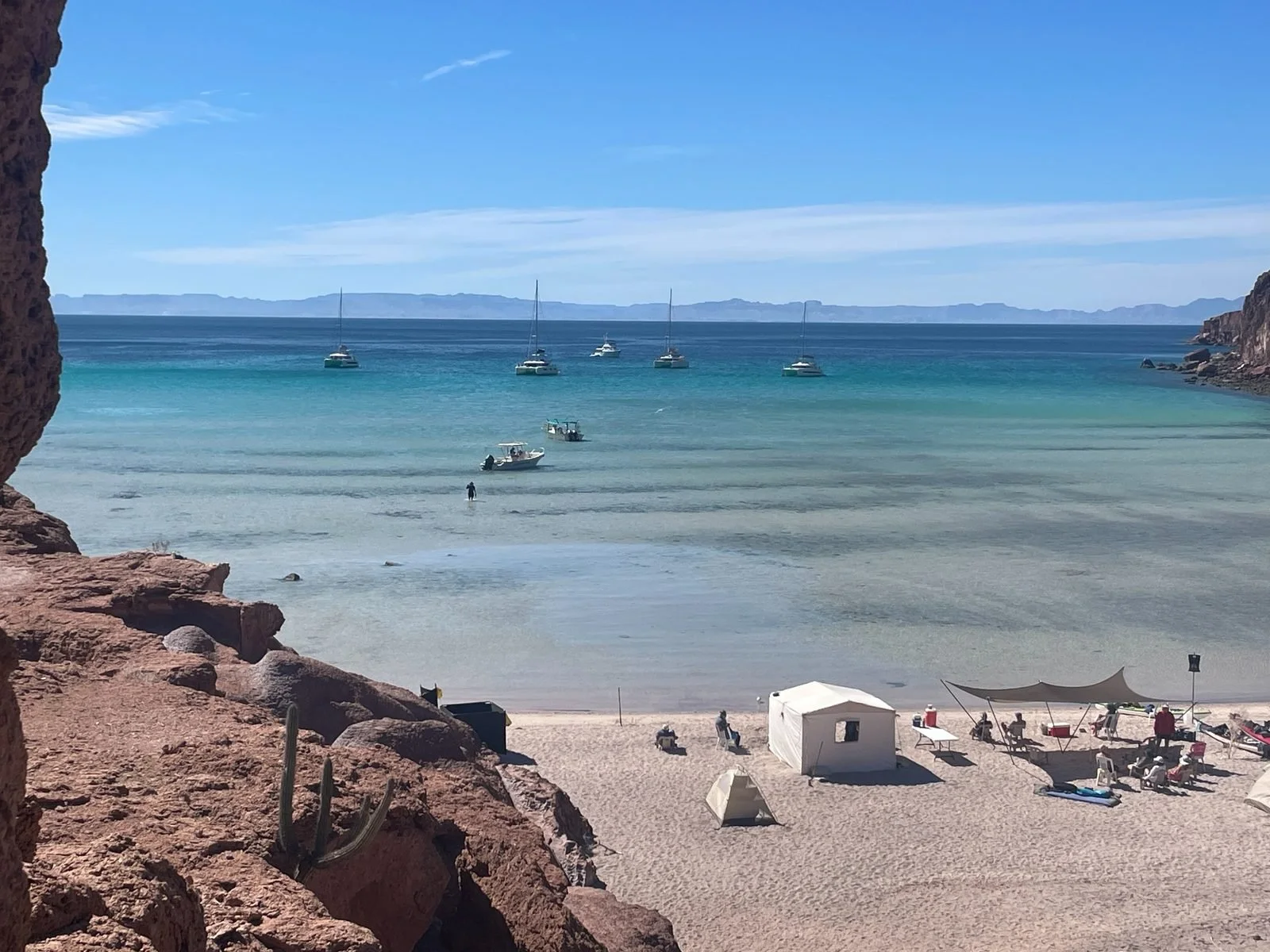 A beach scene with rocks framing the view of the ocean, with several boats anchored near the shore, people on the sand, and a clear blue sky.