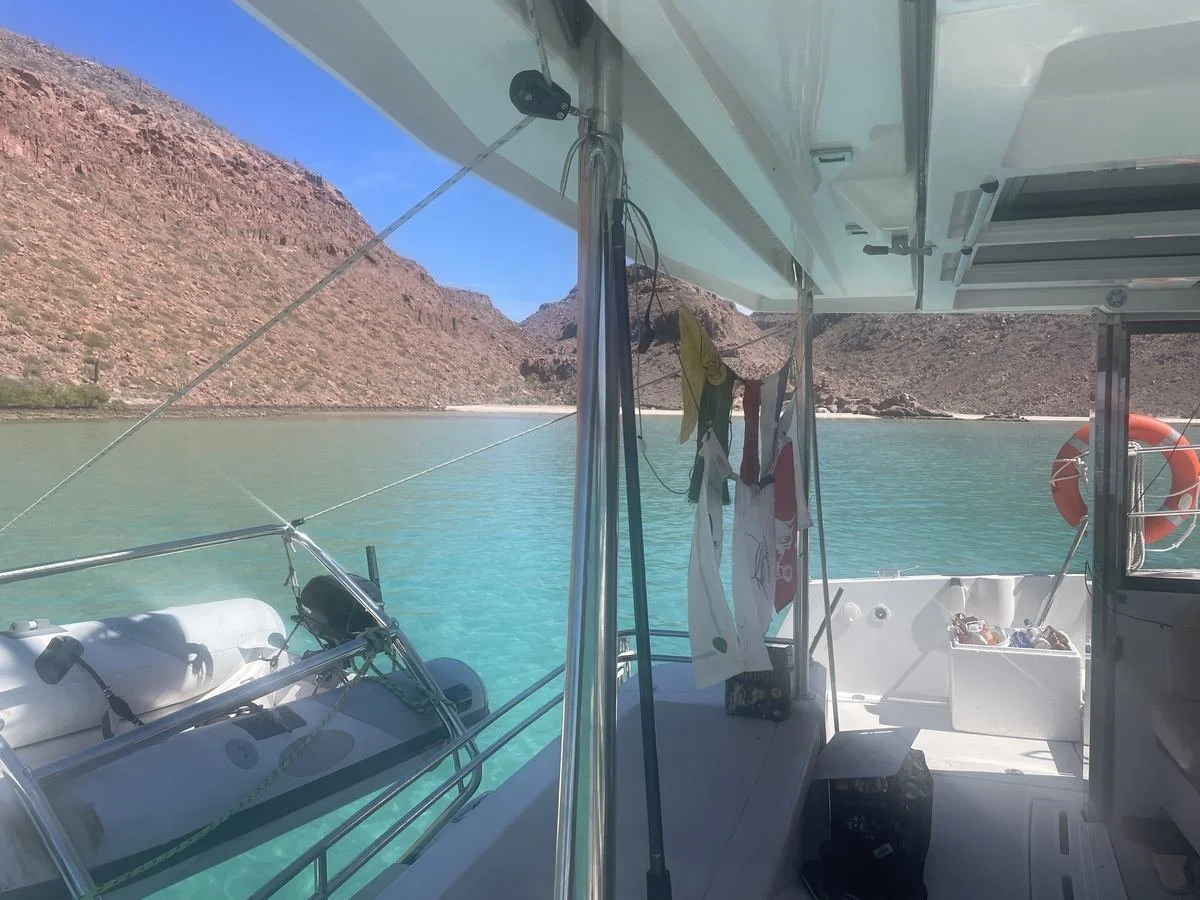 View from a boat looking out at a calm, clear body of water with red rocky hills in the background. The boat has various items hanging and containers visible on deck.