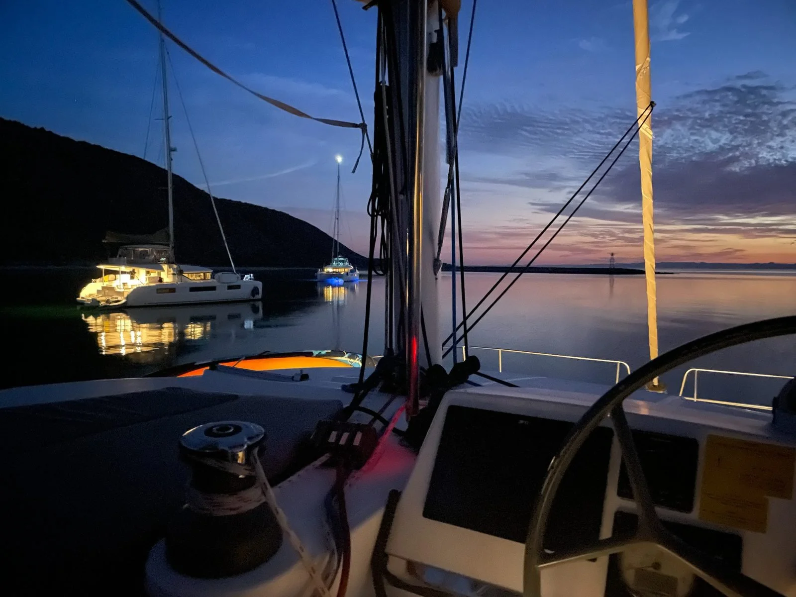 View from a boat at dusk showing calm water with a few anchored boats, a dark hillside on the left, and a colorful sky with clouds and a setting sun on the right.