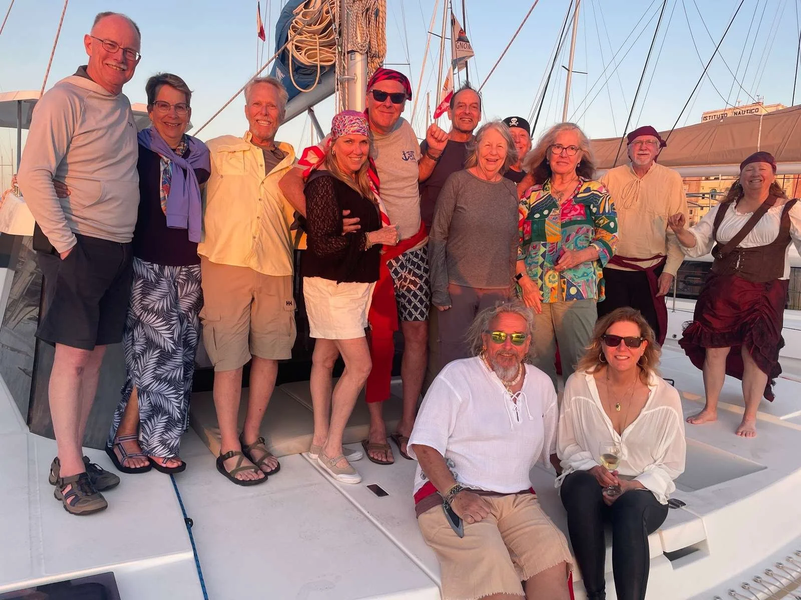 Group of 14 people on a boat posing for a photo at sunset, some sitting and some standing, wearing casual and festive attire, with sailboats and a cityscape in the background.