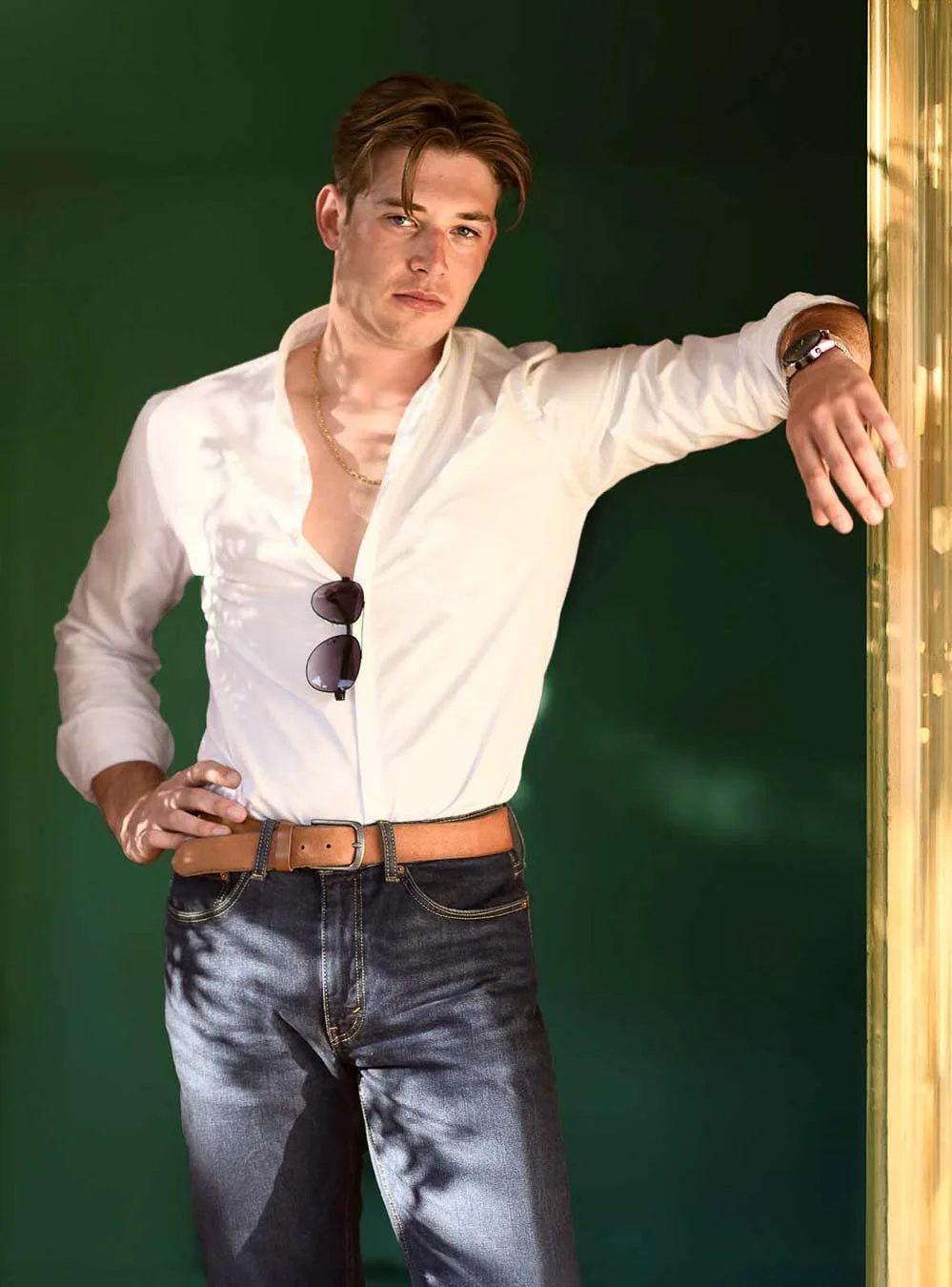 Editorial-style portrait of a young man in a white shirt and jeans against a deep green background
