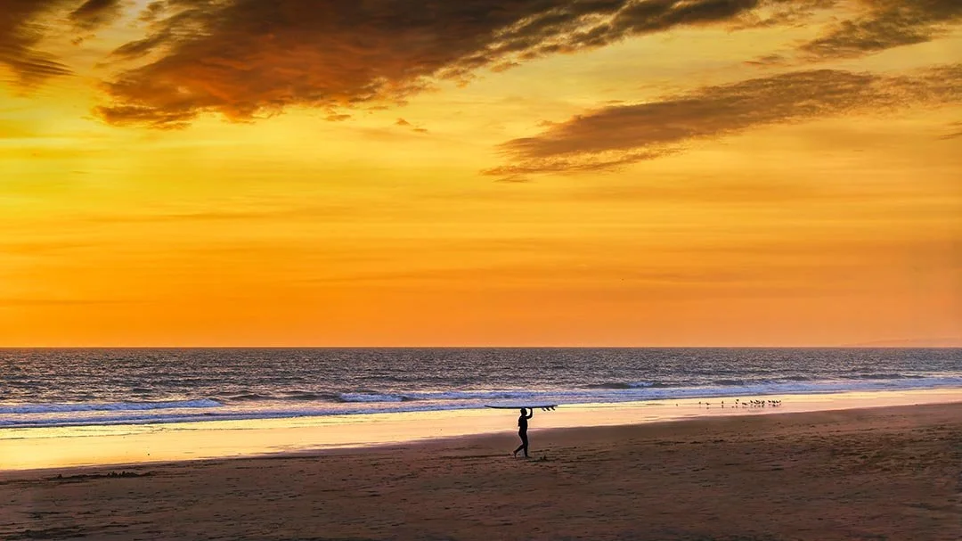 Surfer carrying a board along Falésia Beach beneath a dramatic orange sunset sky
