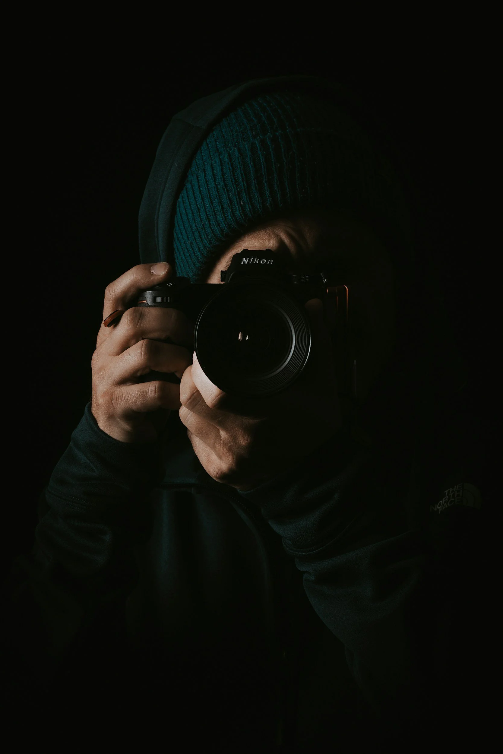 Person in dark clothing and a beanie hat taking a photo with a Nikon camera in a dark environment.