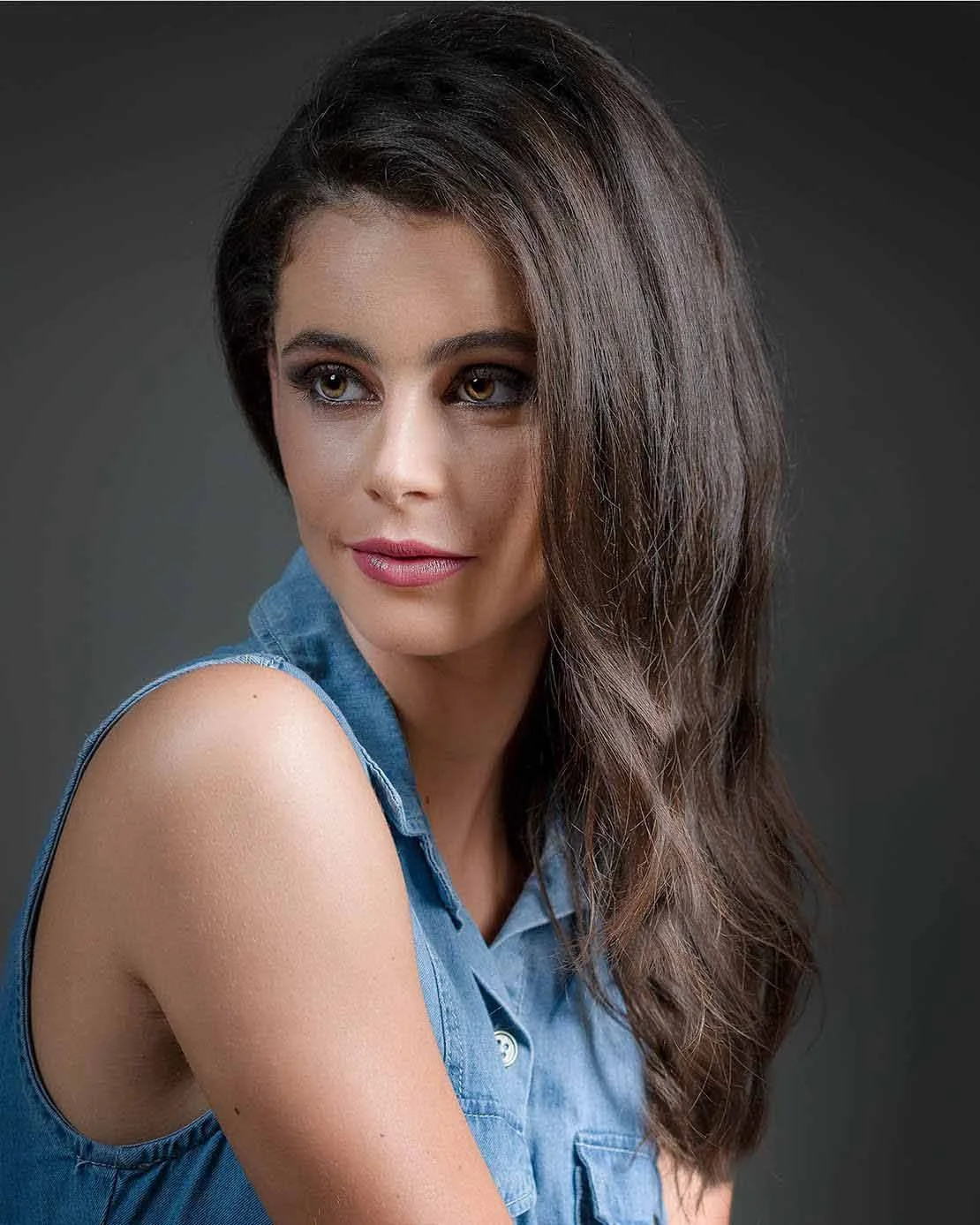 A woman with long, brown, wavy hair and light skin, wearing a sleeveless denim shirt, looking slightly to the side, against a dark background.