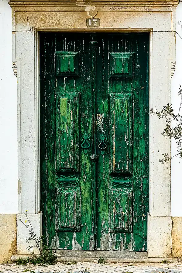 A weathered green wooden door with peeling paint, set in a stone frame, with two old-style door handles and a small architectural feature above the door.