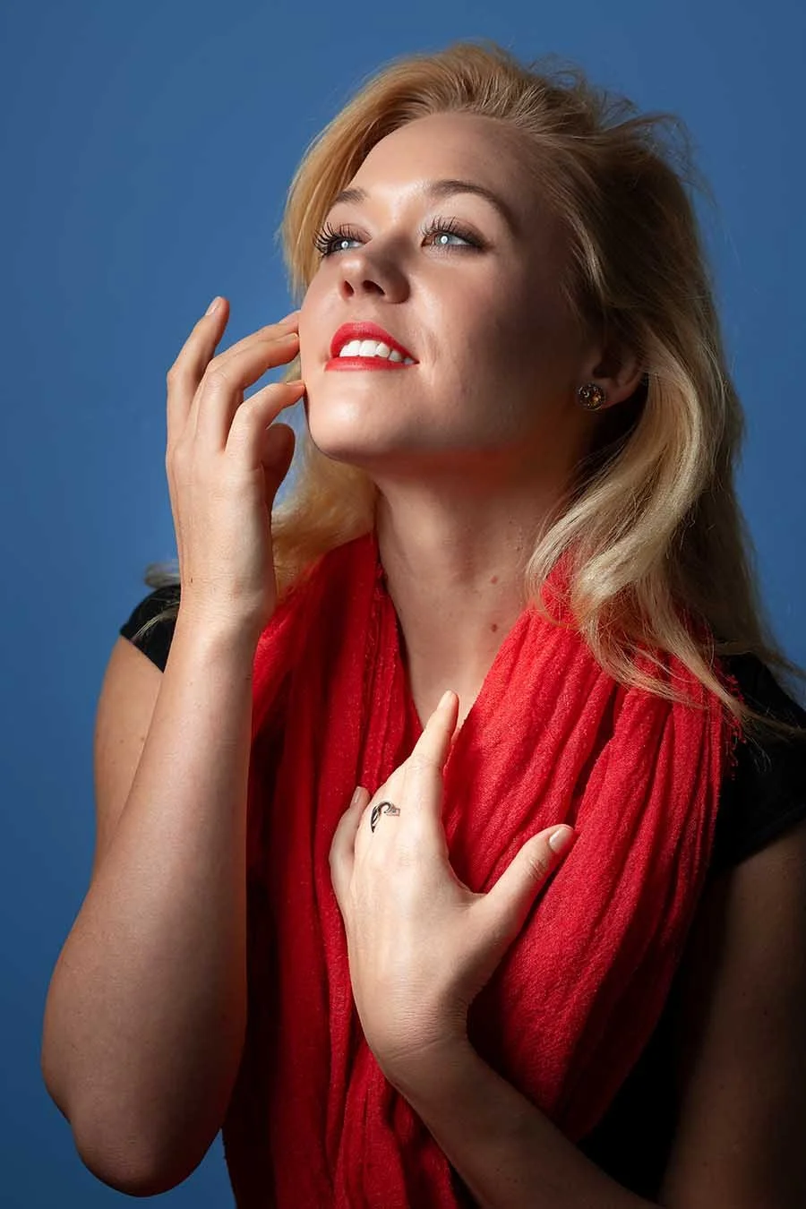 A woman with blonde hair, red lipstick, and a red scarf, posing against a blue background, with her hand near her face and a gentle smile.