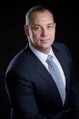 Portrait of a man in a dark suit and light blue tie against a black background.