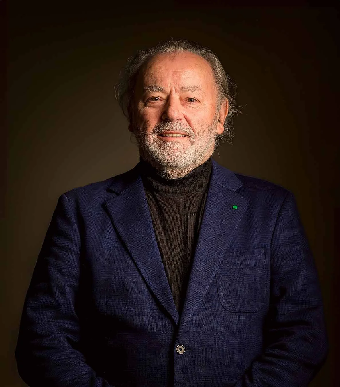 Character portrait of an older man in a blue jacket photographed against a dark studio background