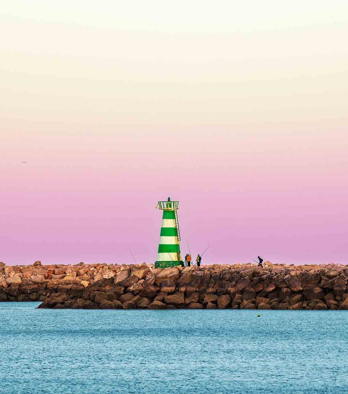 Green Vilamoura harbour lighthouse with anglers beneath a soft pink evening sky