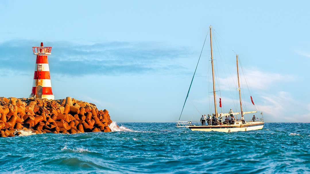 A sailboat sailing near a red and white striped lighthouse on a rocky breakwater with calm blue water and partly cloudy sky in the background.