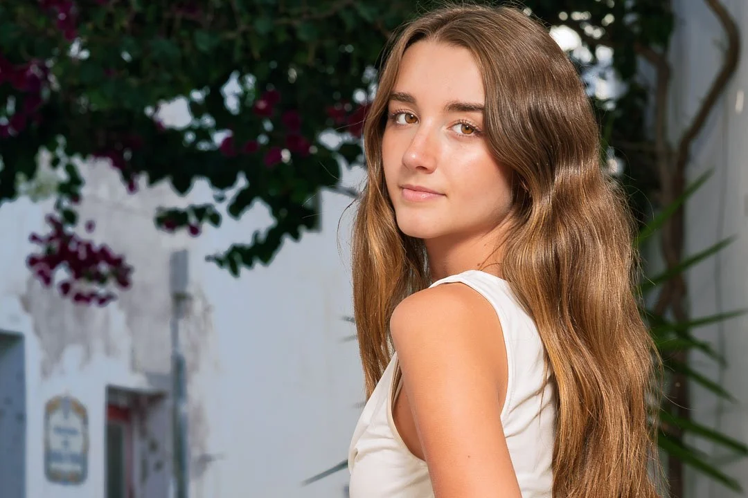 Close-up portrait of a woman looking over her shoulder, with flowers and a whitewashed wall behind her in Albufeira.