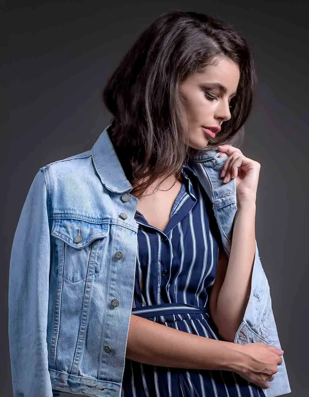 A woman with dark brown, shoulder-length hair wearing a denim jacket over a navy and white striped dress.