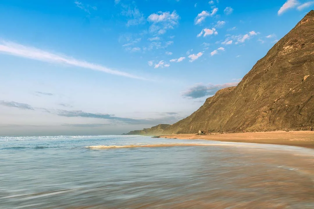 Scenic beach with waves and sandy shore, mountain cliffs on the right, blue sky with clouds.