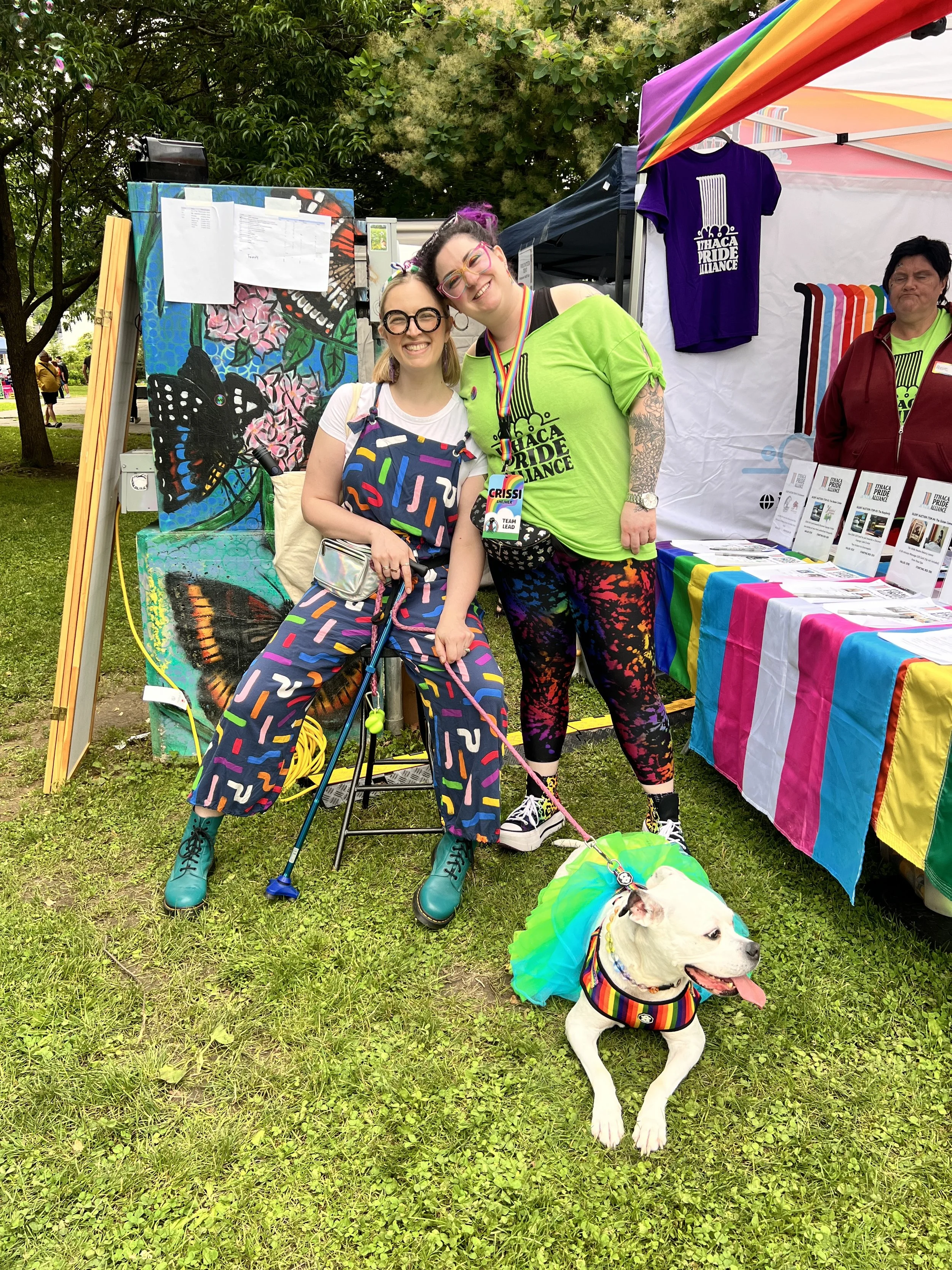 Two people wearing colorful clothing smile, one of them is sitting on a stool with a cane and holding the leash of a dog