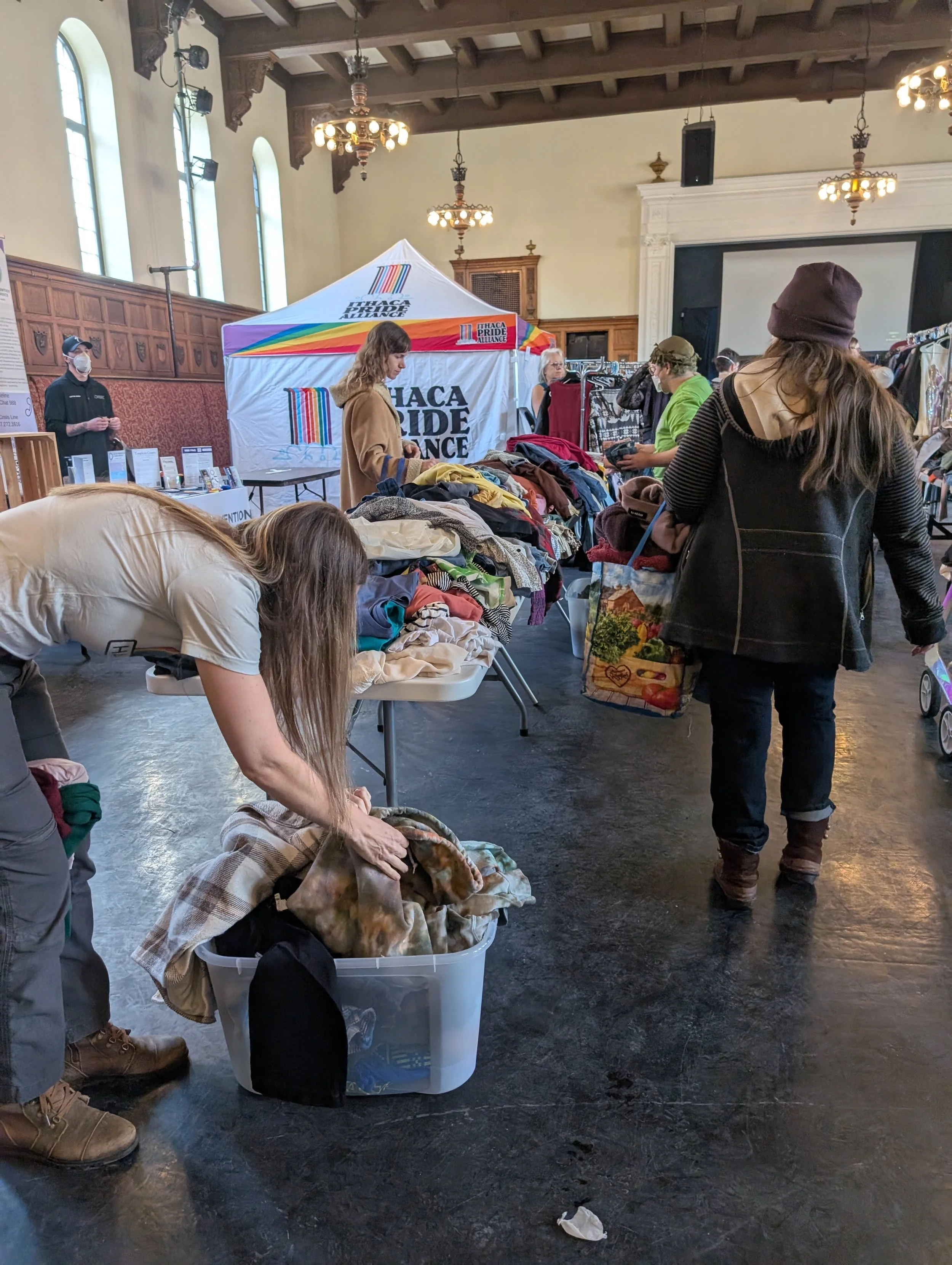 Large piles of clothing are displayed on tables with many people picking up items to