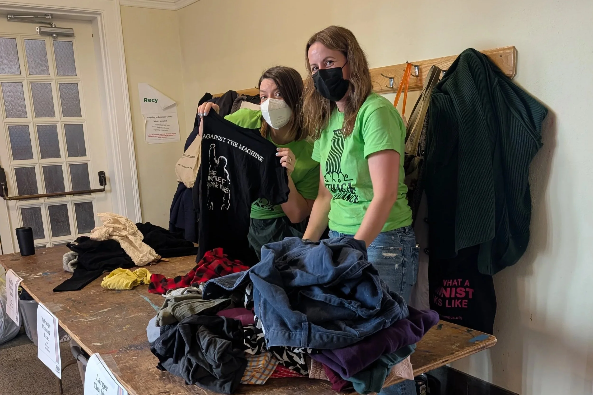 Two people wearing N95 masks pose with clothing being sorted for use at a clothing swap