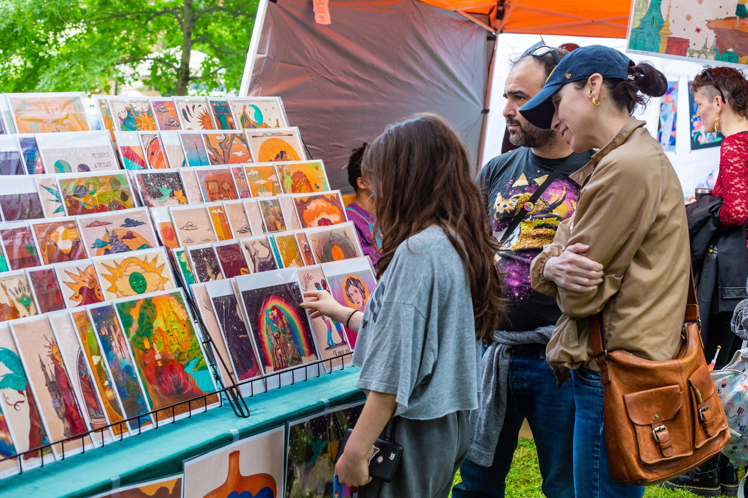 A child and two adults look at a display of printed art outside with a festival happening in the background