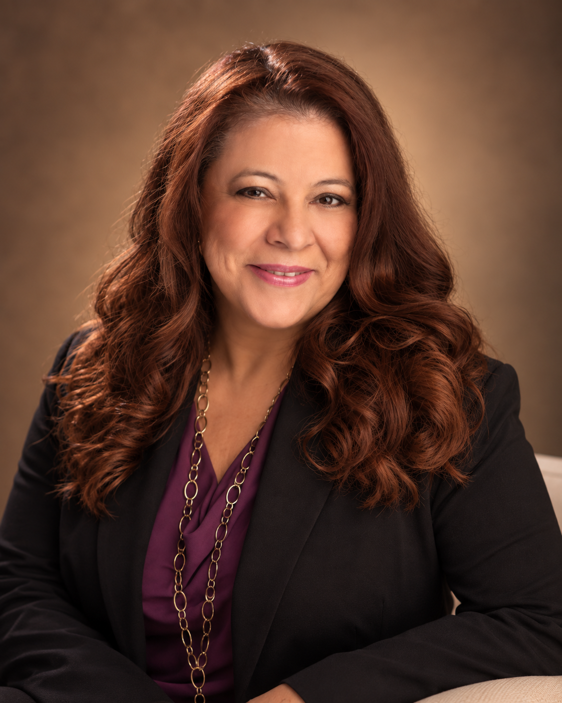 A woman with long, wavy, reddish-brown hair smiling, wearing a black blazer and a purple blouse, with gold jewelry, in a professional portrait setting.