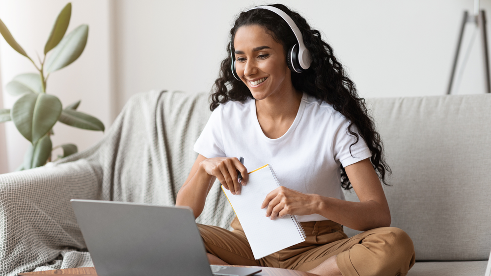 A student smiling on a couch with a laptop, headphones and a notepad