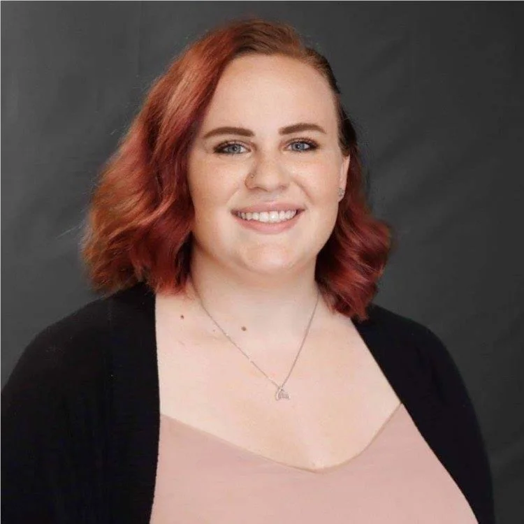 A smiling woman with shoulder-length red hair, blue eyes, and fair skin, wearing a black cardigan over a light pink top and a silver necklace with a small charm, standing against a dark gray background.