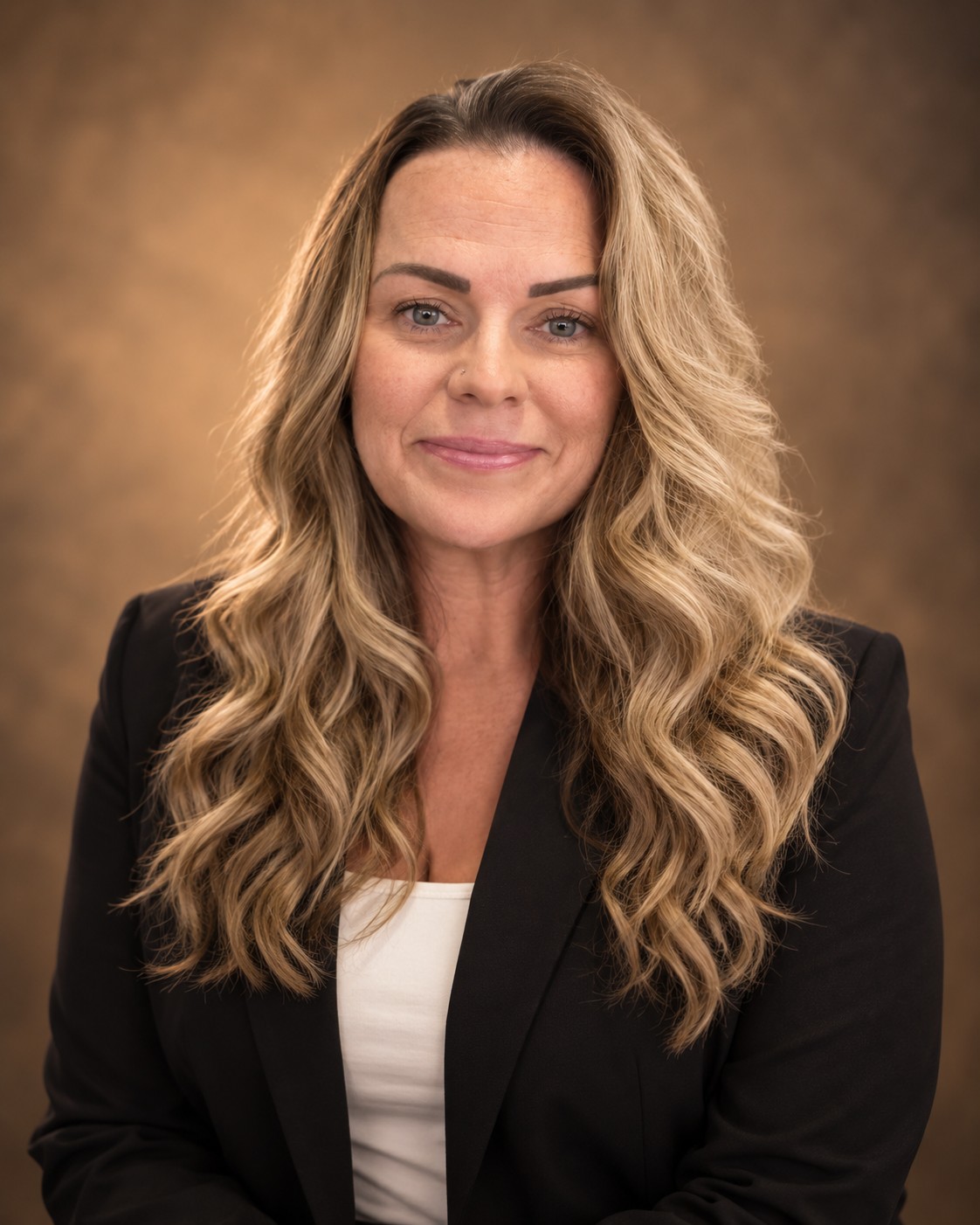 A woman with long, wavy blonde hair, wearing a black blazer and white top, smiling softly against a brown, textured background.