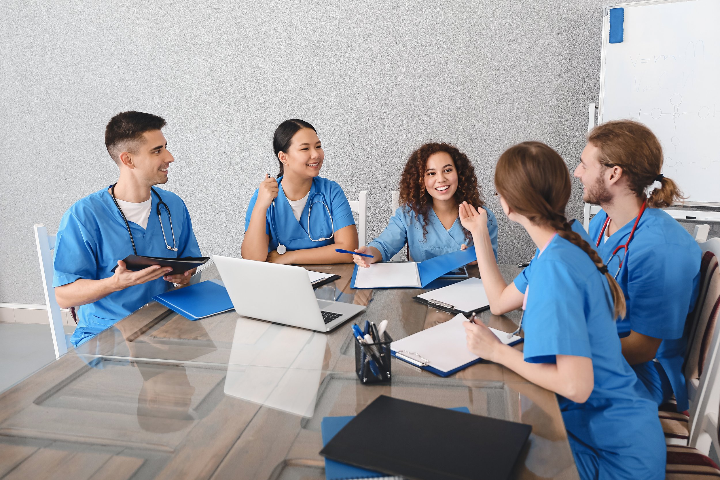 Group of five diverse healthcare professionals in blue scrubs having a meeting in a conference room with notebooks, laptops, and a whiteboard.