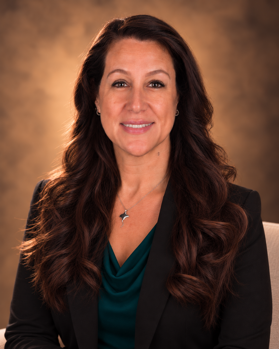 A woman with long, wavy brown hair, wearing a dark blazer and a teal blouse, smiling, with a warm brown background.