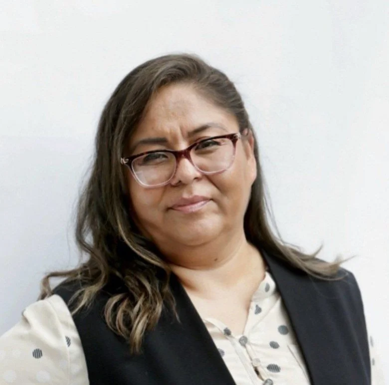 A woman with shoulder-length brown hair, wearing glasses, a light-colored blouse with polka dots, and a dark vest, standing against a white background.