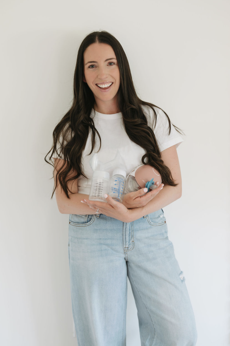 Smiling woman with long dark hair holding baby bottles and a pacifier in front of a plain white wall.