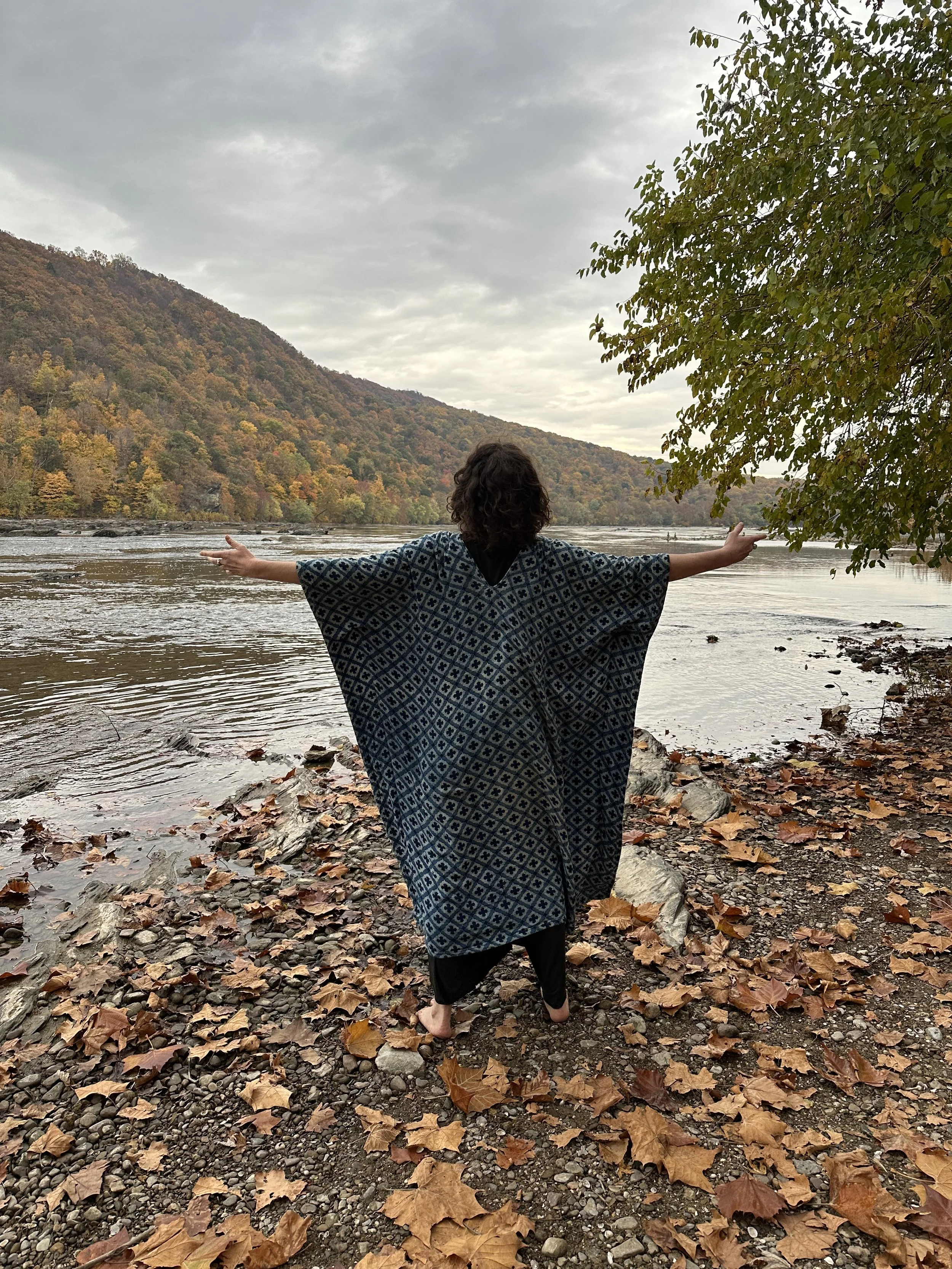 Person in a patterned cloak standing by a river with autumn foliage, arms outstretched, facing hills in the background.