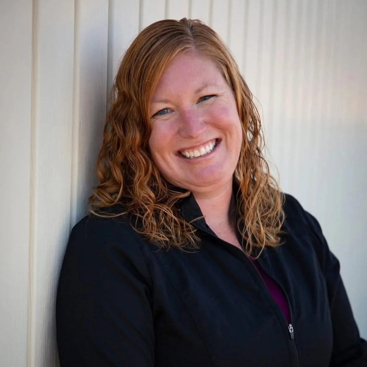 A woman with red curly hair smiling, leaning against a cream-colored wall, wearing a black jacket.